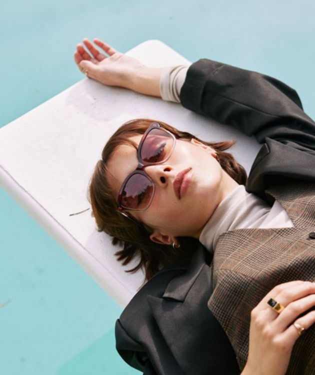 Frau mit modischer, roter Sonnenbrille auf einem Pool-Sprungbrett liegend.